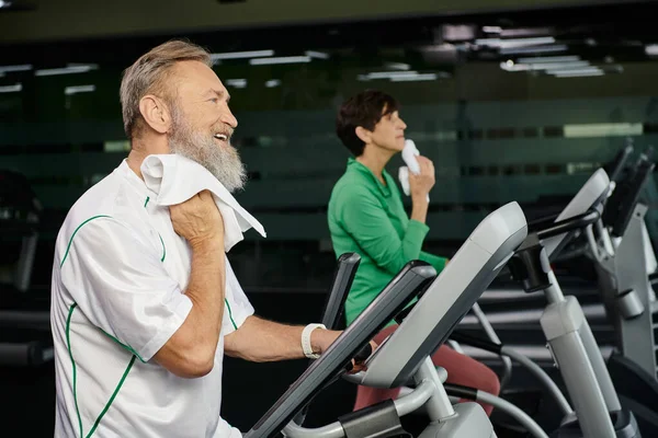 senior and bearded man wiping sweat with towel, blurred woman on background, exercising in gym