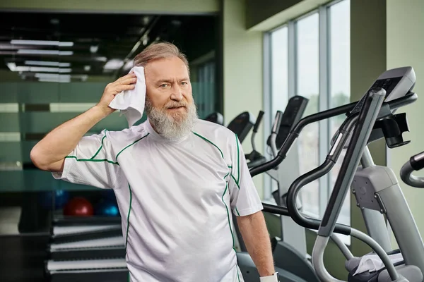 elderly man with beard wiping sweat with towel after working out in gym, looking at camera, sport