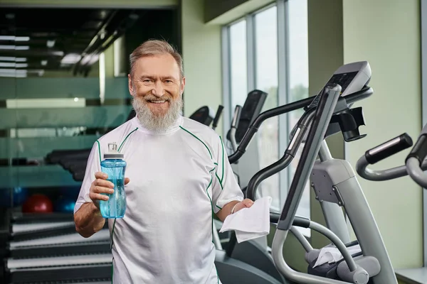 happy elderly man holding sports bottle with water and smiling after working out in gym sport