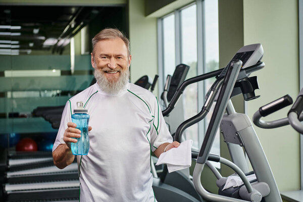 happy elderly man holding sports bottle with water and smiling after working out in gym sport