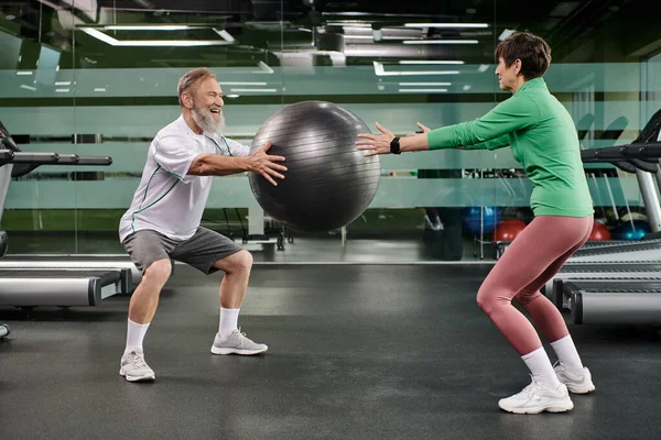 happy elderly man and woman working out with fitness ball in gym, active seniors, couple sport
