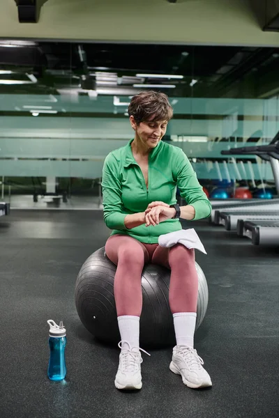happy elderly woman sitting on fitness ball, looking at fitness watch after workout, sports bottle