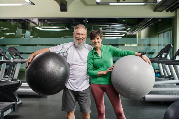 sporty elderly couple, cheerful man and woman holding fitness balls, active seniors in gym