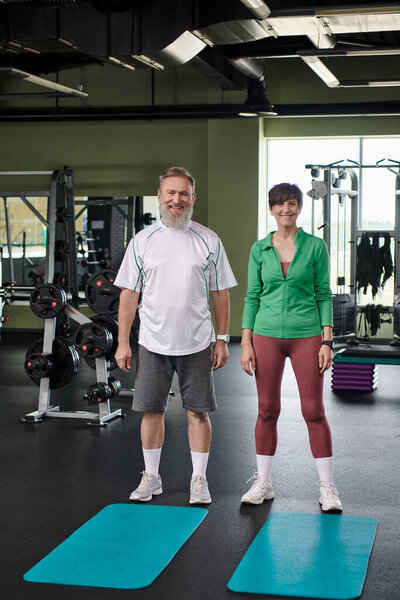 happy elderly couple standing near fitness mats, looking at camera, full length, active seniors