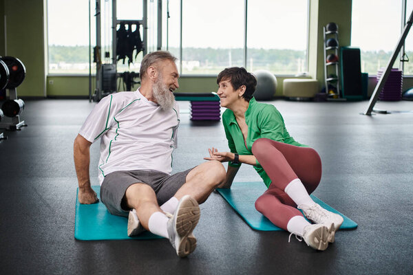elderly man and woman looking at each other, active seniors exercising on fitness mats in gym