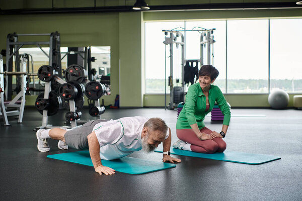 пожилой мужчина с бородой делает доску, счастливая женщина упражнения в тренажерном зале, фитнес коврики, активные пожилые люди