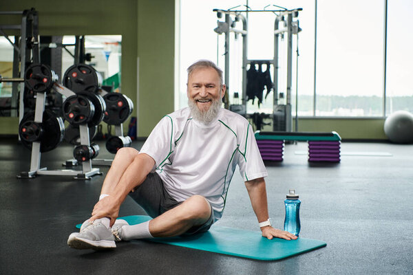 веселый пожилой человек с бородой сидит на тренажерном коврике возле спортивной бутылки с водой, активный старший