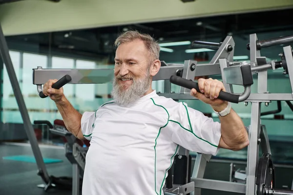 strong and elderly man with beard working out on exercise machine, athletic and healthy, gym