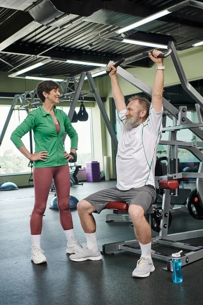 strong and elderly man with beard working out on exercise machine near happy woman, gym, active