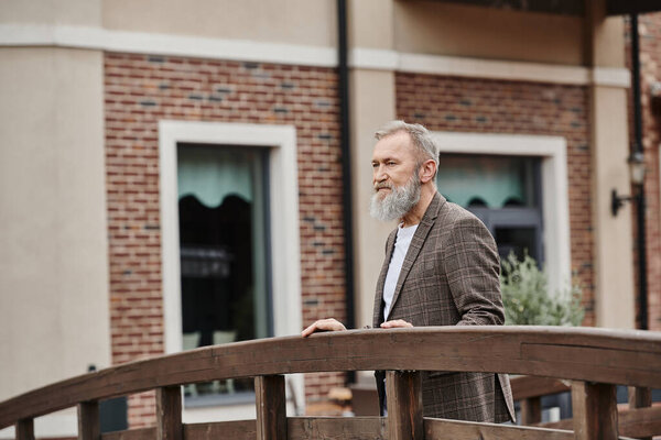 bearded elderly man with grey hair standing on wooden bridge, looking away, thinking, urban backdrop