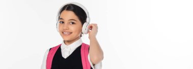 Müzik, kablosuz kulaklıklı neşeli okul kızı beyaz afişte izole edilmiş kameraya bakıyor.