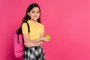 happy schoolgirl with backpack holding green fresh apple isolated on pink, vibrant backdrop clipart