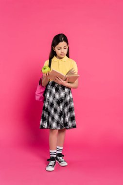 full length, brunette schoolgirl holding green apple and reading book on pink background, backpack clipart