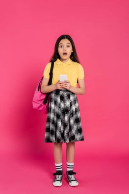 Şaşkın kız, elinde akıllı telefon tutan kız öğrenci ve pembe arka plandaki kameraya canlı bir şekilde bakıyor.