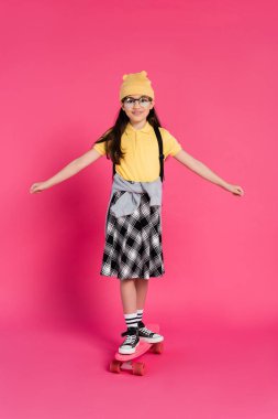 happy schoolgirl in beanie hat and glasses riding penny board on pink background, stylish look clipart