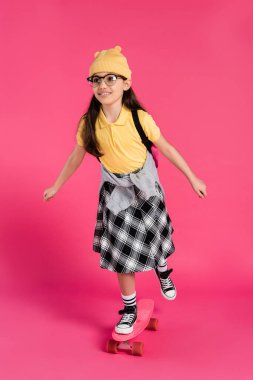 cheerful schoolgirl in beanie hat and glasses riding penny board on pink background, stylish look clipart