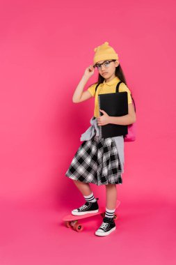 schoolgirl in beanie hat and glasses holding notebooks, standing near penny board on pink background clipart