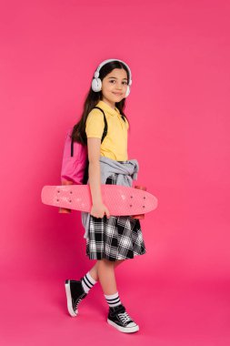 Kablosuz kulaklıklar takan mutlu okul kızı dersten sonra tahta üzerinde pembe arka planda duruyor.