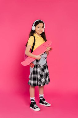 Kablosuz kulaklıklı neşeli kız dersten sonra elinde bozuk para tahtasıyla pembe arka planda duruyor.