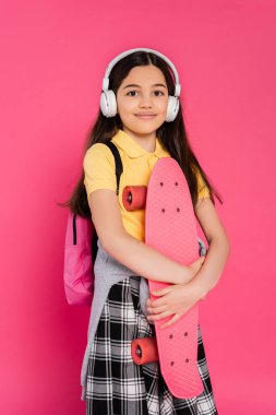 Kablosuz kulaklık takan pozitif kız dersten sonra tahta üzerinde pembe arka planda duruyor.