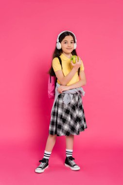 Kablosuz kulaklıklı gülümseyen kız pembe arka planda yeşil elma tutuyor, sırt çantalı çocuk.