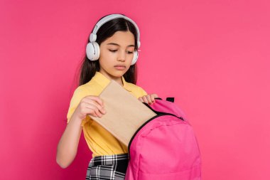 Kablosuz kulaklıklı esmer kız. Sırt çantasına kitap koyuyor. Pembe arka plan, öğrenci.