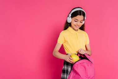 Kablosuz kulaklıklı mutlu kız öğrenci sırt çantasına elma koyuyor, pembe arka plan, öğrenci