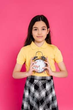 brunette schoolgirl holding alarm clock isolated on pink, looking at camera, back to school clipart