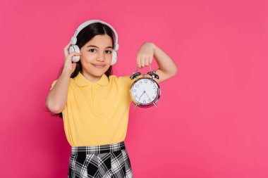 happy schoolgirl in wireless headphones holding vintage alarm clock isolated on pink, student life clipart