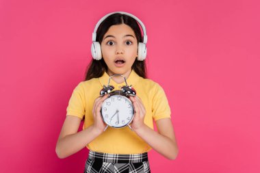 shocked schoolgirl in wireless headphones holding vintage alarm clock isolated on pink, student life clipart