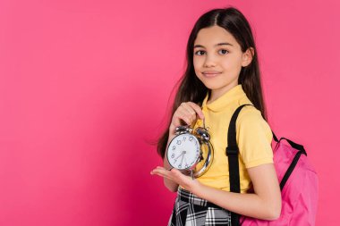 happy brunette schoolgirl standing with backpack and holding vintage alarm clock isolated on pink clipart