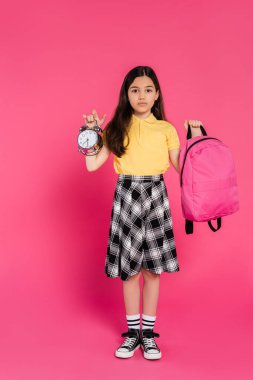 full length, brunette schoolgirl standing with backpack and holding vintage alarm clock on pink clipart