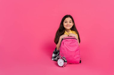 cheerful brunette schoolgirl sitting with backpack near vintage alarm clock on pink background clipart