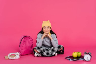 happy schoolgirl in beanie hat sitting near notebooks, headphones, apples, backpack and alarm clock clipart