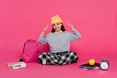 happy schoolgirl in beanie hat showing v sign, notebooks, headphones, apple, backpack, alarm clock clipart