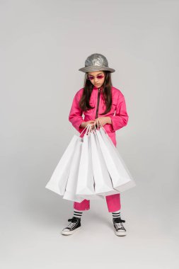 Pembe elbiseli şık bir kız ve gri arka planda alışveriş çantaları taşıyan panama şapkalı, uzun boylu.