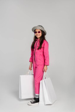 Şık kıyafetli, güneş gözlüklü ve panama şapkalı, gri arka planda alışveriş torbaları taşıyan neşeli bir kız.