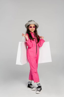 Şık giyinmiş, güneş gözlüklü ve panama şapkalı, gri arka planda alışveriş torbaları taşıyan gülümseyen bir kız.