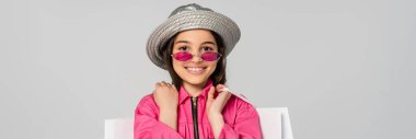 positive girl in stylish outfit, sunglasses and panama hat posing with shopping bags on grey, banner clipart