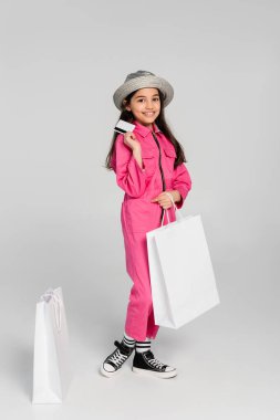 Moda kıyafetli ve panama şapkalı gülümseyen kız alışveriş çantalarının yanında poz veriyor, gri üzerinde kredi kartı tutuyor.