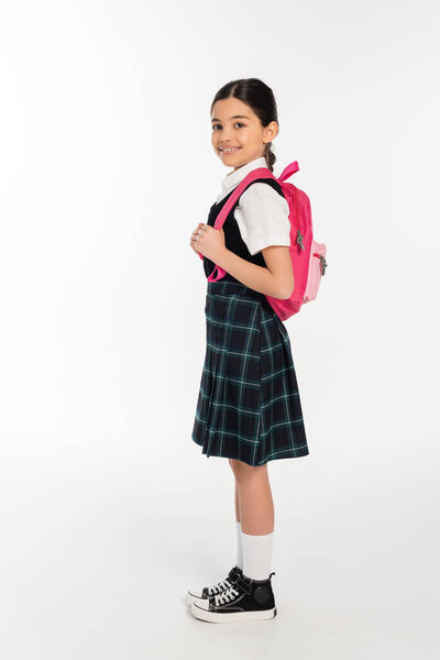 full length, happy schoolgirl standing in uniform with backpack on white, ready for new school year