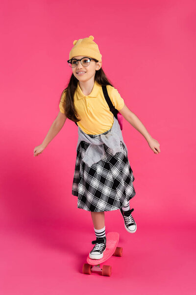 cheerful schoolgirl in beanie hat and glasses riding penny board on pink background, stylish look
