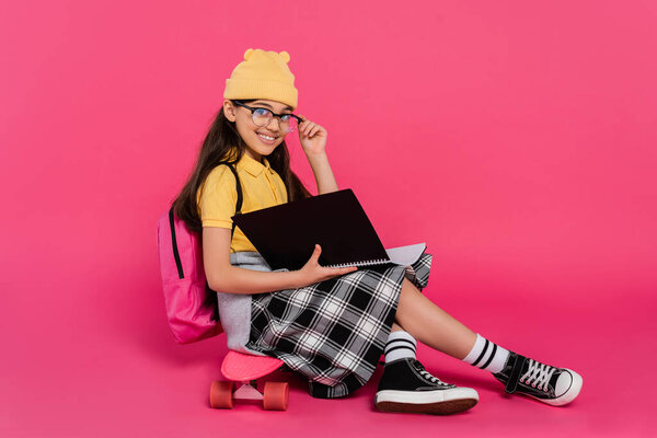 cheerful schoolgirl in beanie hat and glasses sitting on penny board, pink background, notebooks