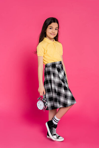 happy schoolgirl holding alarm clock on pink background, student looking at camera, full length