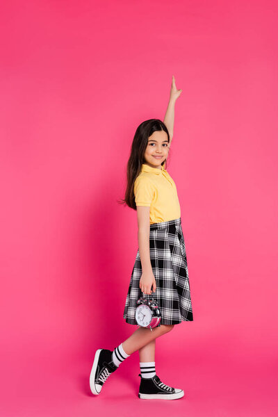 happy schoolgirl holding alarm clock on pink background, looking at camera, pointing up, full length