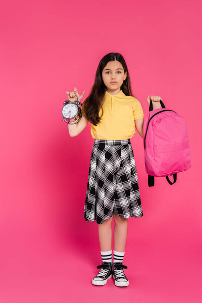 full length, brunette schoolgirl standing with backpack and holding vintage alarm clock on pink