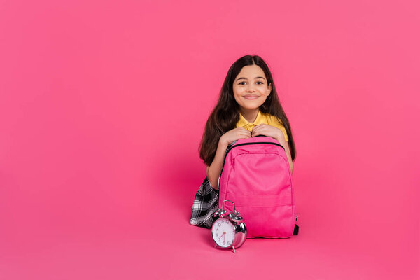 cheerful brunette schoolgirl sitting with backpack near vintage alarm clock on pink background