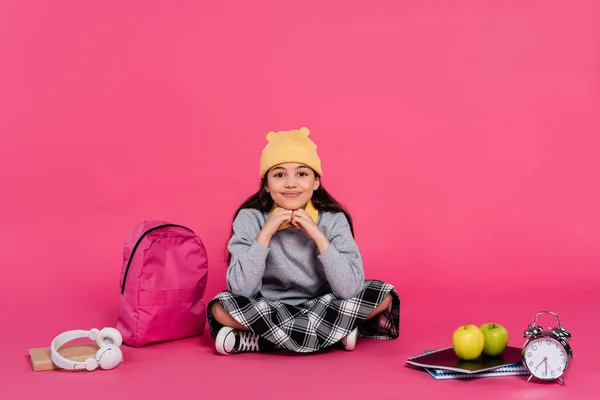happy schoolgirl in beanie hat sitting near notebooks, headphones, apples, backpack and alarm clock