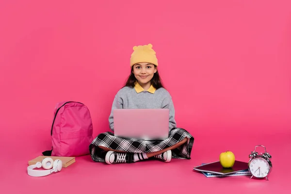 happy schoolgirl in beanie hat using laptop, sitting near headphones, apple, backpack, alarm clock