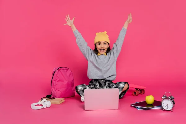 cheerful girl in beanie hat using laptop, sitting near headphones, apple, backpack, alarm clock
